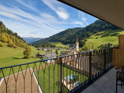 Balkonaussicht auf grünes Tal mit Dorf und Bergen unter blauem Himmel.