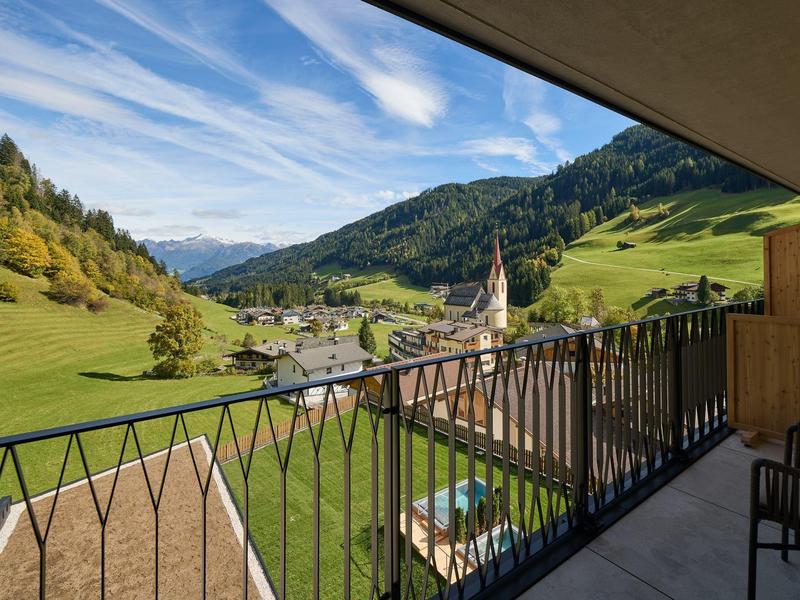 Blick von einem Balkon auf ein grünes Tal mit Bergen, Dorf und blauem Himmel.