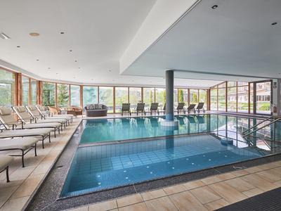 Moderner Innenpool mit Liegestühlen, großen Fenstern und hellem Fliesenboden in einem Wellnessbereich.