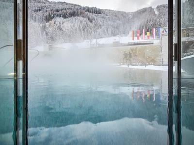 Blick aus einem warmen Innenpool auf verschneite Berge und bewölkten Himmel draußen.
