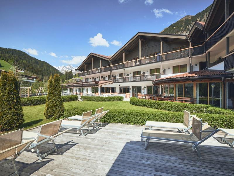 Moderner Hotelbau mit Terrasse, Sonnenliegen, grüner Wiese und Bergkulisse unter blauem Himmel.