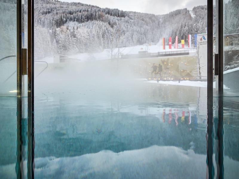Blick aus einem warmen Innenpool auf verschneite Berge und bewölkten Himmel draußen.