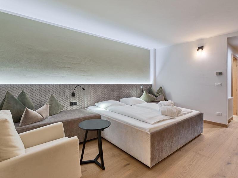 Modernes Schlafzimmer mit Doppelbett, beige Sessel, Holzboden und warmweißer Beleuchtung.