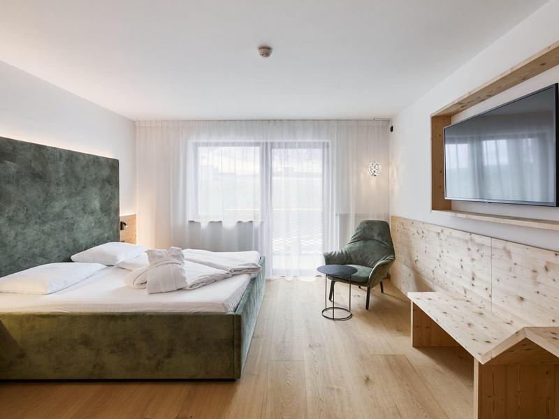 Helles Hotelzimmer mit Doppelbett, Holzfußboden, grünem Sessel und Wand-TV.