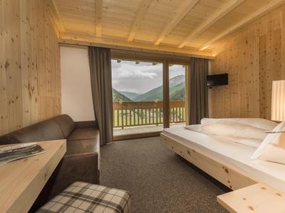 Gemütliches Holz-Zimmer mit großem Bett, Sofa, Balkon und Bergblick, warmes Licht und Teppichboden.
