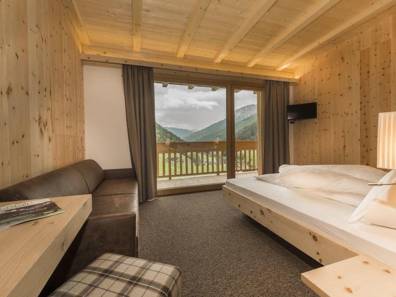 Gemütliches Holz-Zimmer mit großem Bett, Sofa, Balkon und Bergblick, warmes Licht und Teppichboden.