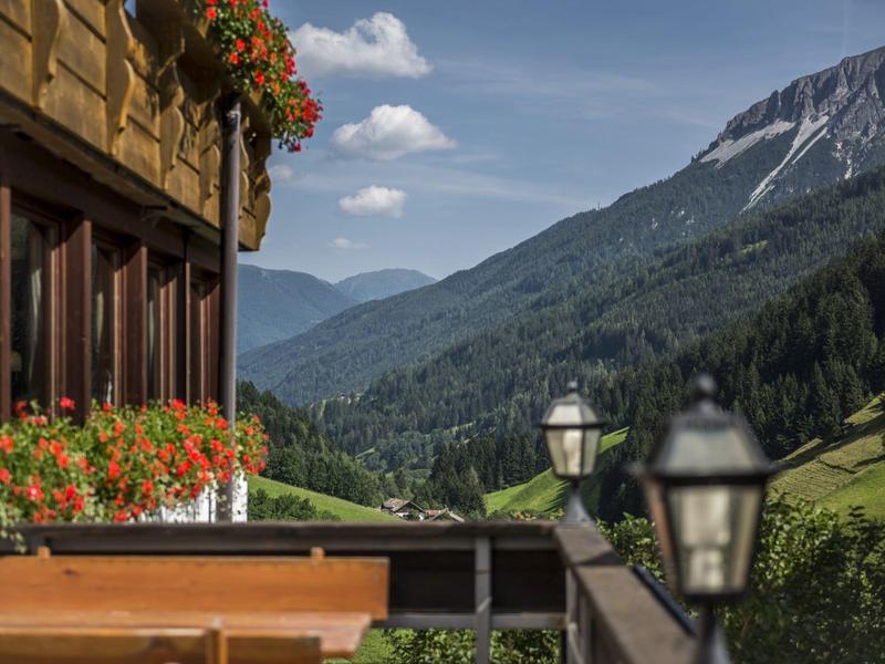 Berglandschaft mit Tal, grünen Wiesen, Holzhaus mit roten Blumen und klar blauem Himmel.