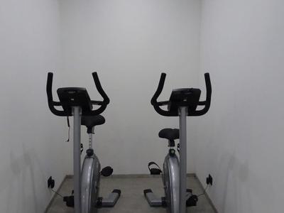 Zwei stationäre Fahrräder stehen nebeneinander in einem hellen, engen Raum mit weiß gestrichenen Wänden.