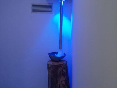 Kunstinstallation mit Baumstamm, Wasserschale und blau beleuchtetem Fallrohr an weißer Wand.