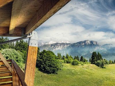 Mountain landscape with green valley, trees, and snowy peaks, viewed from a wooden terrace.