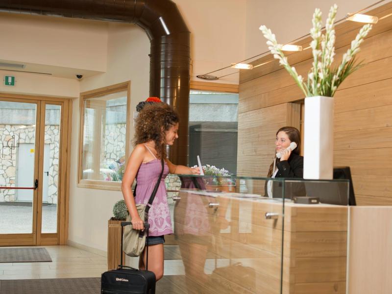 Eine Frau checkt an der Hotelrezeption ein, mit Rollkoffer und Rucksack.