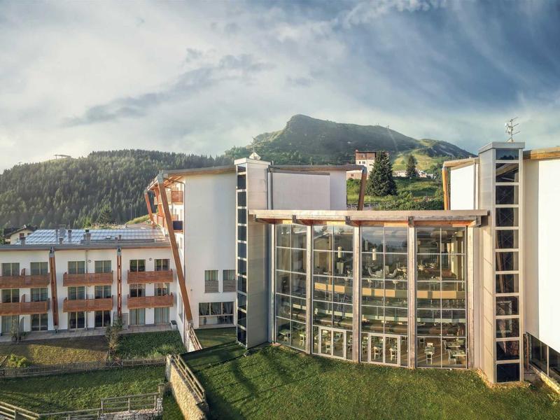 Modernes Hotel mit großer Glasfassade vor bewaldeten Bergen und blauem Himmel.