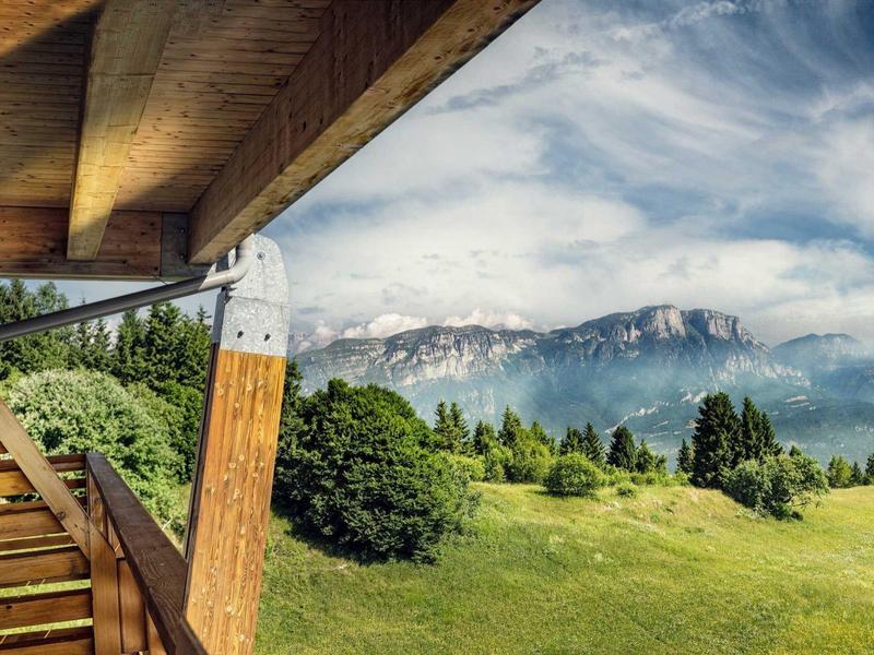 Blick von einer Holzbalkonveranda auf grüne Wiesen und bewaldete Berge unter wolkigem Himmel