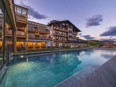 Moderner Hotelkomplex mit beleuchtetem Außenpool bei Dämmerung und Bergblick im Hintergrund.