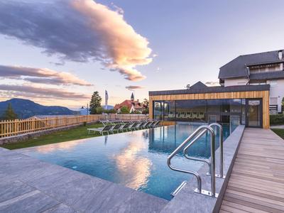 Moderner Außenpool mit Glasgeländer auf Holzdeck, Berglandschaft und Himmel mit Wolken im Hintergrund.