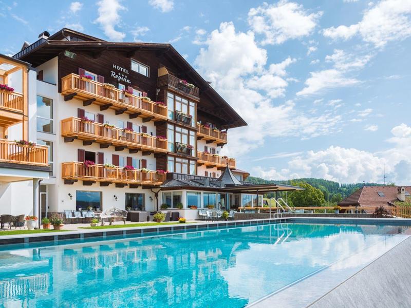 Großes Holzhaus mit Balkonen neben einem klaren blauen Schwimmbecken unter blauem Himmel.