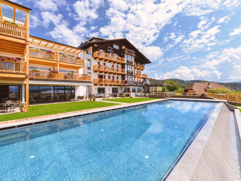 Moderner Außenpool vor einem großen, traditionellen Hotel mit Holzdetails und blauen Himmel.
