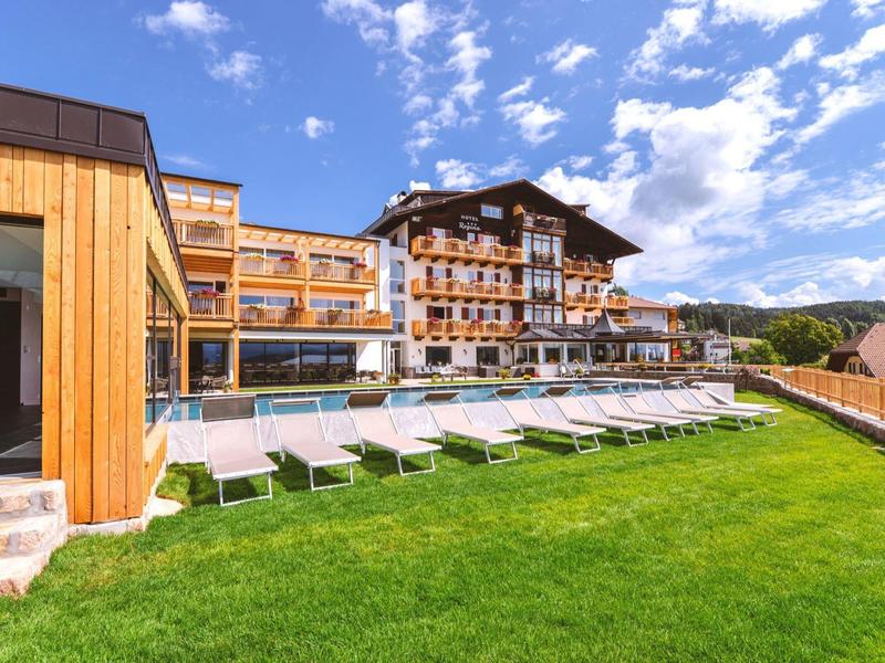 Großes Hotel mit Holzfassade, Poolbereich und Liegen auf grünem Rasen unter blauem Himmel.