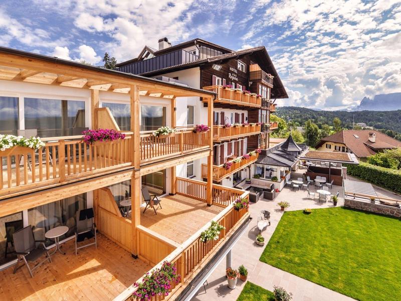 Großes Holzhaus mit Balkonen, Blumen und einer grünen Rasenfläche bei sonnigem Himmel.