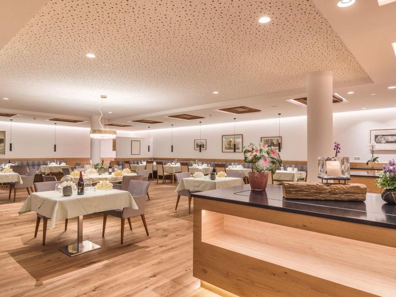 Großer, heller Speisesaal mit Holzboden, weißen Tischen und Stühlen, dekorativen Blumenarrangements.