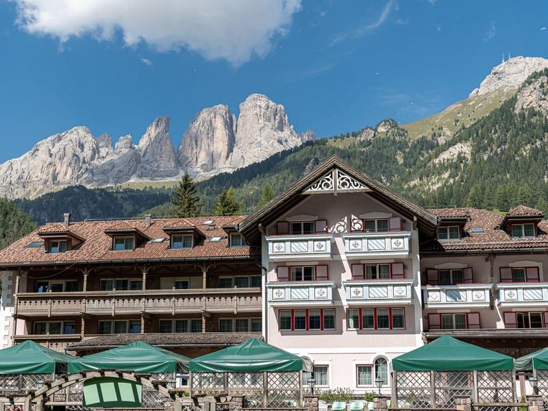 Großes Alpenhotel mit braunem Dach und weißer Fassade vor bewaldeten Bergen und blauem Himmel.