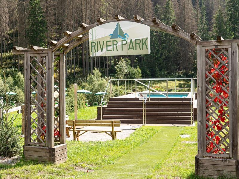Parkeingang mit Holzbogen, Bänken, Rasen und Wald im Hintergrund unter sonnigem Himmel.