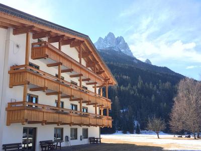 Holzhaus mit Balkonen neben verschneitem Feld und bewaldetem Berg im Hintergrund unter blauem Himmel.