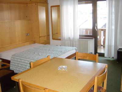 Einfach eingerichtetes Zimmer mit Doppelbett, Holztisch mit vier Stühlen und Fenster mit Gardinen.