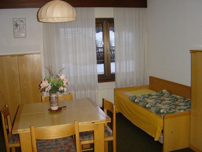 Kleiner Raum mit Holzmöbeln, Tisch, Stühlen, Bett mit Blumenbettwäsche und Fenster mit weißen Gardinen.