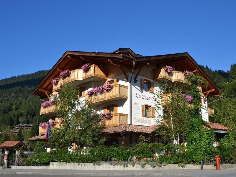 Großes Alpenhotel mit Holzverzierungen, Blumen auf Balkonen, grünem Busch und blauem Himmel.
