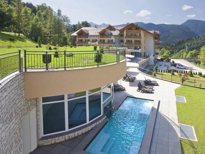 Hotel mit Pool, grüner Wiese, Holzchalets und Bergen im Hintergrund unter blauem Himmel.
