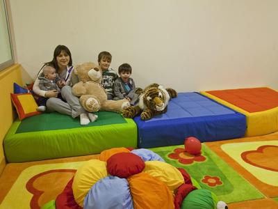 Drei Kinder sitzen auf buntem Polstermöbel mit Plüschtieren in einem hellen Raum.