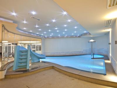 Innenraum mit beleuchtetem blauen Schwimmbecken und kleiner Rutsche, helle Wände, moderne Decke.