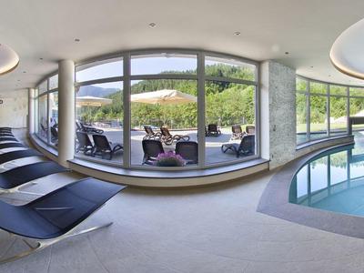 Moderner Wellnessbereich mit Glasfront, Pool, grauen Liegen und Blick auf grüne Landschaft.