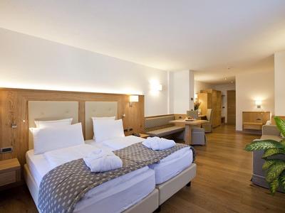 Helles Hotelzimmer mit zwei Einzelbetten, Holzboden, Schreibtisch, Sitzbereich und Grünpflanze.