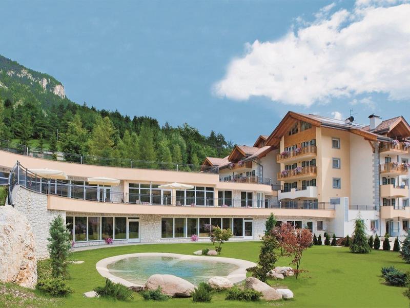Großes Hotelgebäude mit mehreren Stockwerken, Swimmingpool und Garten vor bewaldetem Hügel.