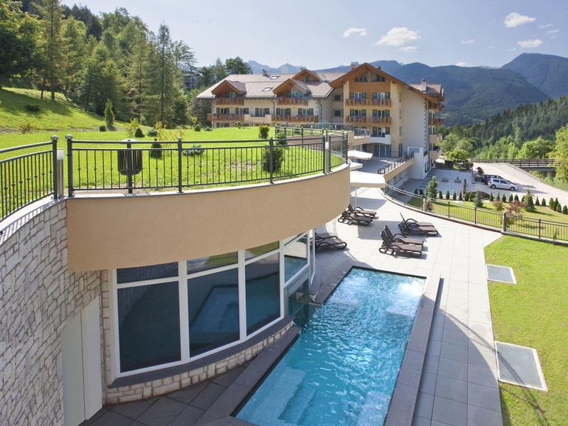 Hotel mit Pool, grüner Wiese, Holzchalets und Bergen im Hintergrund unter blauem Himmel.