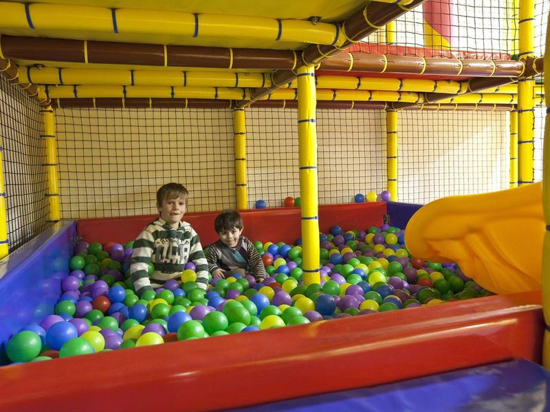 Kinder spielen in einem bunten Bällebad mit gelben Netzstreben und einer gelben Rutsche.