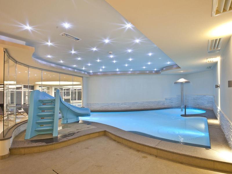 Innenraum mit beleuchtetem blauen Schwimmbecken und kleiner Rutsche, helle Wände, moderne Decke.