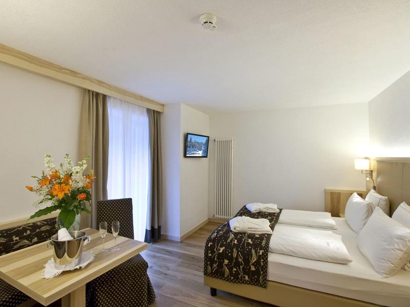 Helles Hotelzimmer mit Doppelbett, Tisch mit Blumen und Fernseher an der Wand.