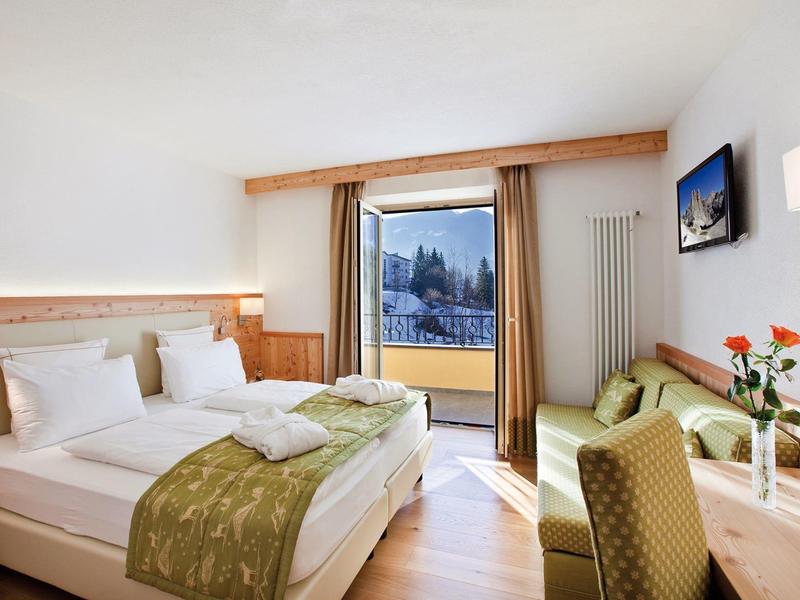 Helles Hotelzimmer mit Doppelbett, Sessel, Tisch, Balkon und Blick auf verschneite Berge.