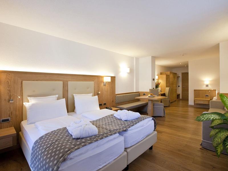 Helles Hotelzimmer mit zwei Einzelbetten, Holzboden, Schreibtisch, Sitzbereich und Grünpflanze.