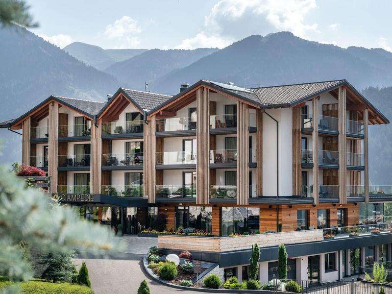 Moderner Alpenhotelkomplex mit Balkonen vor bewaldeten Bergen unter blauem Himmel.