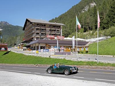 Ein grünes Cabrio fährt vor einem großen Holzgebäude in einer bergigen Landschaft mit grünen Wäldern.