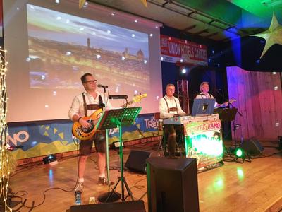 Drei Musiker in traditioneller Tracht spielen auf einer Bühne mit bunten Lichtern und Leinwand im Hintergrund.