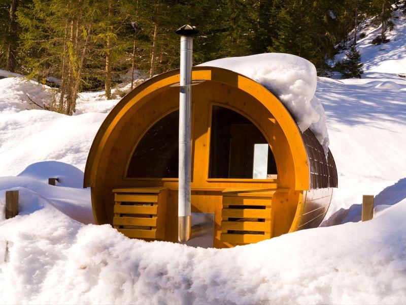 Rundes Holzhaus mit Fenster und Ofen im Schnee vor Waldrand bei Sonnenschein.