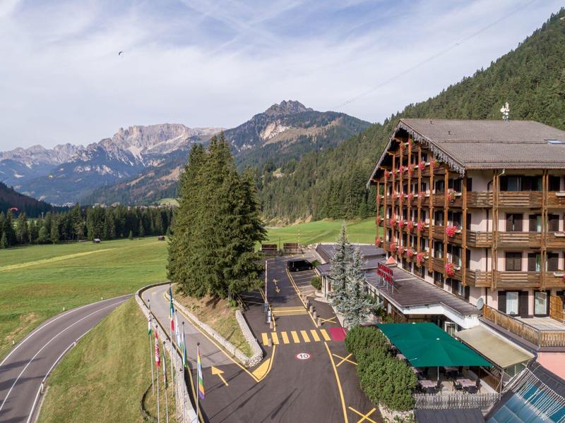 Berglandschaft mit grünem Tal, großer Hotelanlage, Straße und bewaldeten Hängen unter blauem Himmel.