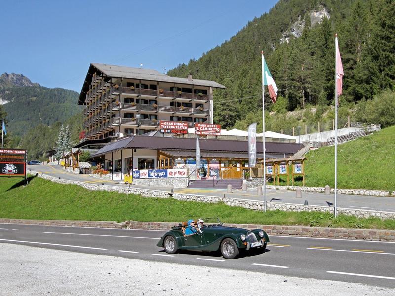 Ein grünes Cabrio fährt vor einem großen Holzgebäude in einer bergigen Landschaft mit grünen Wäldern.