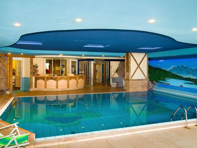 Innenraum mit großem rechteckigem Schwimmbecken, Holztheke, Landschaftswandbild und Liegestühlen.