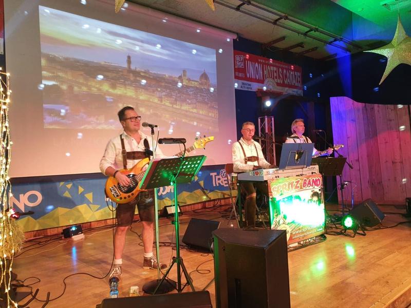 Band in Trachten auf Bühne, bunte Beleuchtung, Leinwand mit Landschaft im Hintergrund.
